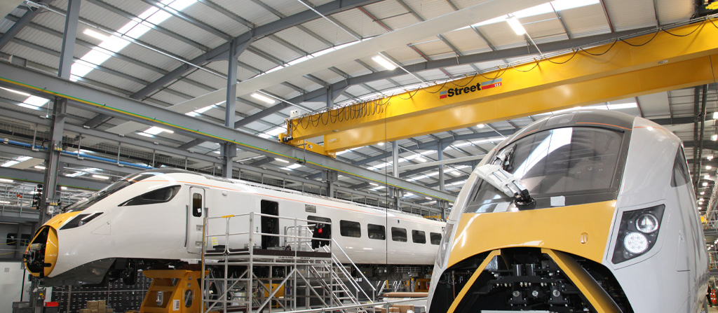 Two trains in a factory with a yellow double girder crane above
