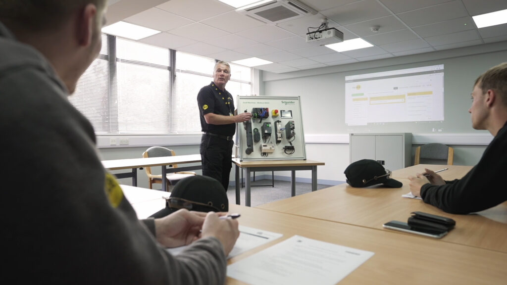 A man standing next to a board with electrical components, teaching some staff members.